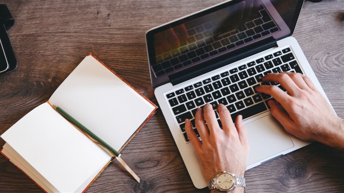 hands typing on a laptop, with an open notebook
