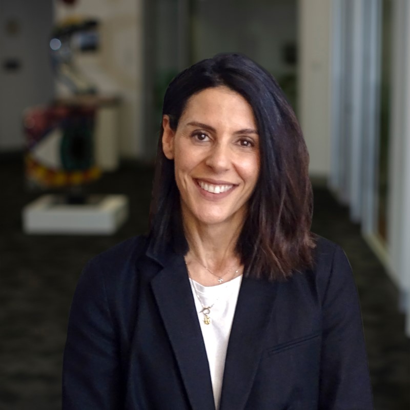 Photo of Rosa Zouzoulas. Rosa is wearing a white shirt and black blazer and is standing in the foyer of the Nillumbik Council building.