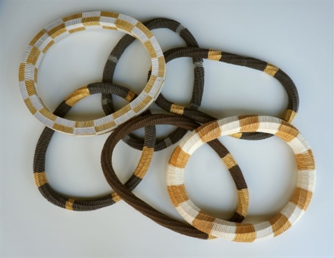 Visual description: Six woven necklaces overlapping each other on a white background. The necklaces have blocks of brown, gold, white and black colours.