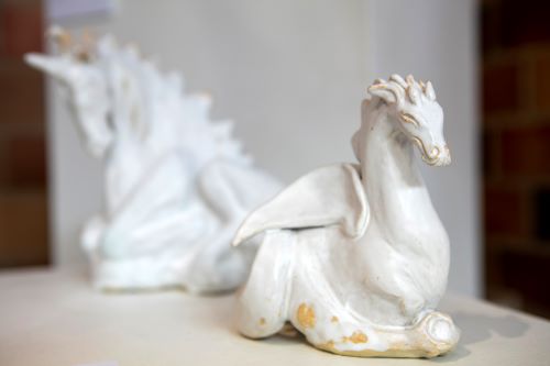 Visual description: Two ceramic creatures that appear to be white unicorns. The creature in the background is blurred in the photo, both creatures are seated and resting.