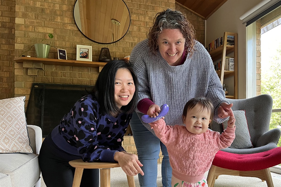 Two women are in a loungeroom supporting a young toddler to stand up