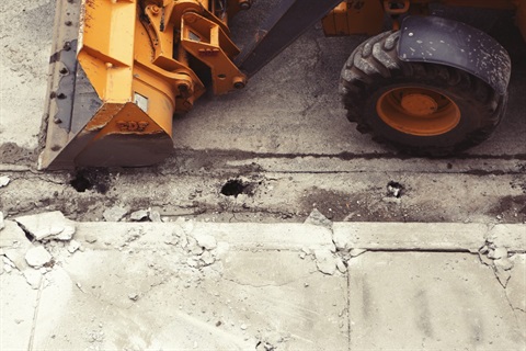 Photo of a large excavator on a road next to a foothpath. The curb of the footpath is damaged.
