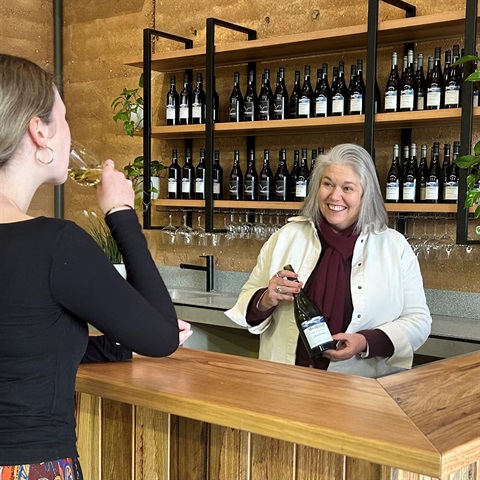 A lady behind a bar holding a bottle of wine smiling at a customer drinking wine