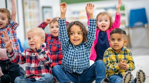 Photo of kindergarten children sitting on the ground with their hands in the air