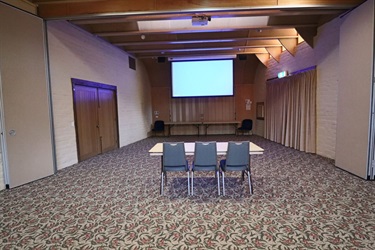 A large carpeted room with a small table in the middle facing toward the end of the room where there is a projector screen lit on the wall