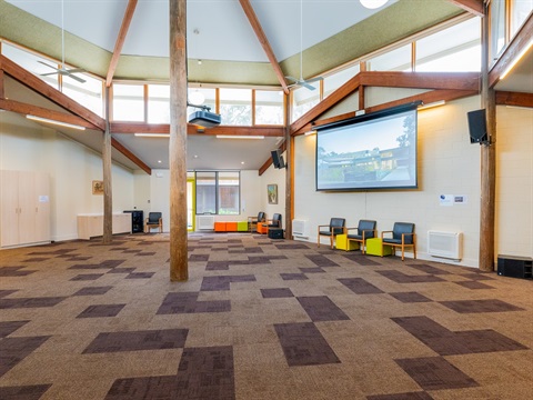 A large carpeted room has windows high around the roofline and a large beam in the middle of the room reaching up to a central ceiling point