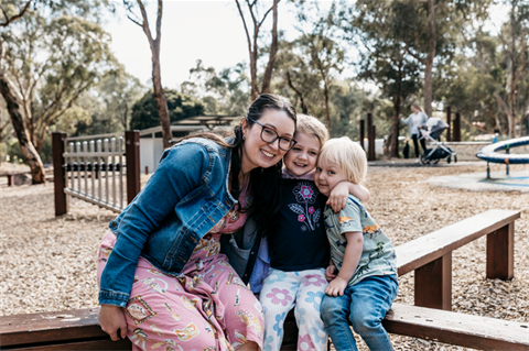A photo of a mother and two children sitting on a bench in a playground. They are smiling and hugging.