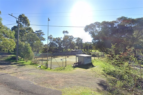 Two tennis courts surrounded by chain link fencing in a rural setting. The tennis court surface is worn and covered in dirt.