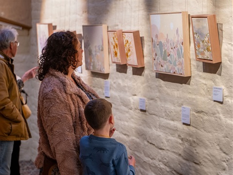 A photo of visitors looking at small landscape paintings on a mudbrick wall