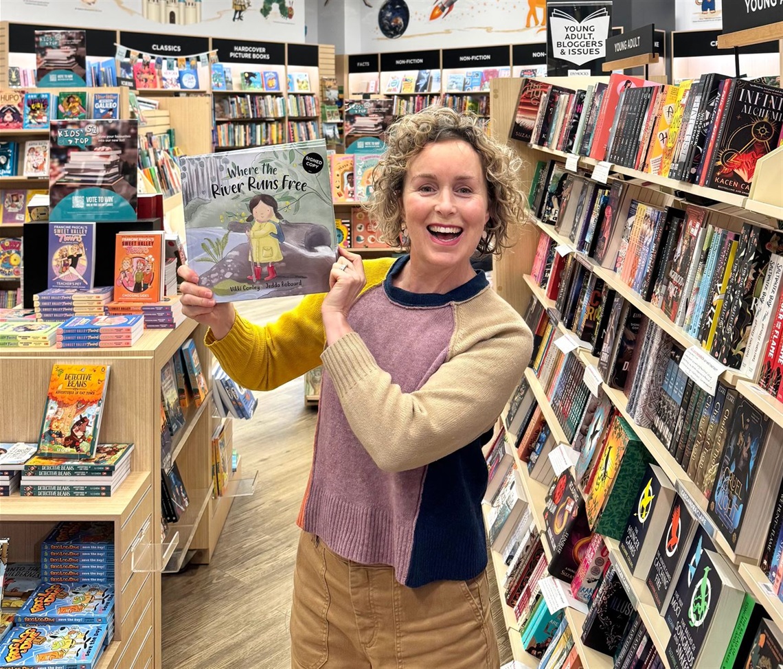 Author Vikki Conley stands in a bookstore smiling and holding a copy of her new book, Where the River Runs Free