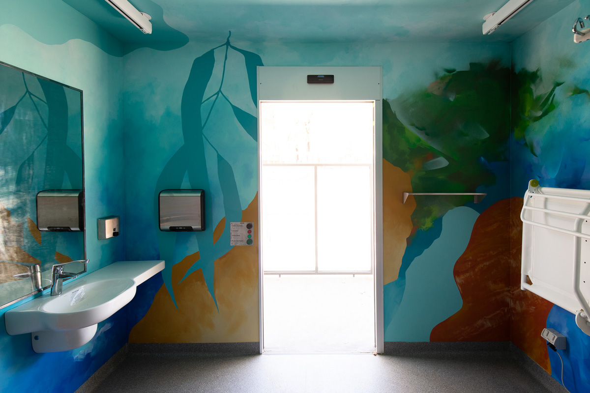 A spacious public bathroom with walls covered in calm colourful artwork, and a sunlit door in the centre of the end wall