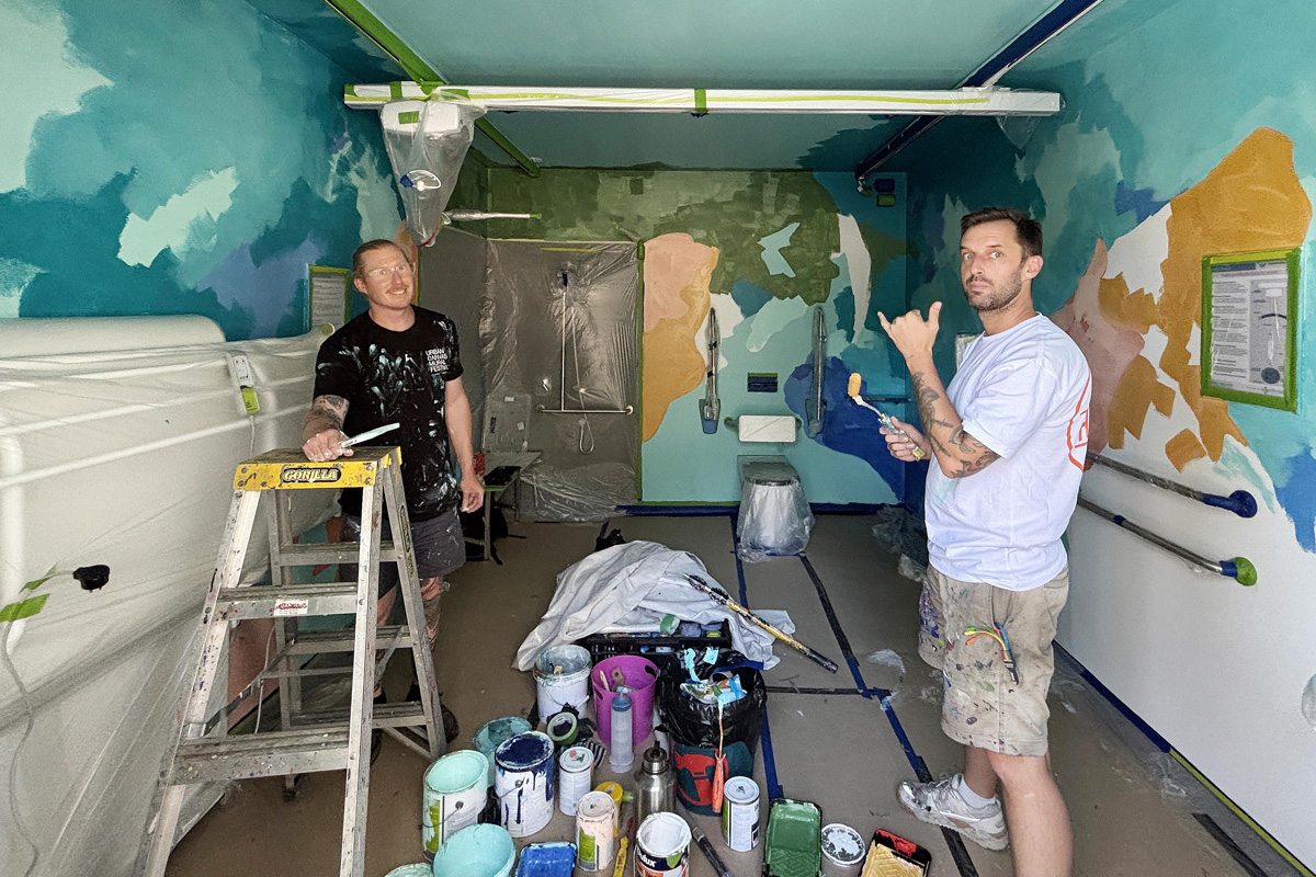 Two men in work gear stand in a public bathroom, there are numerous paint cans on the floor