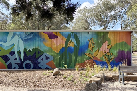 A public building in a park with a long wall covered in a painting of native foliage in blues and oranges