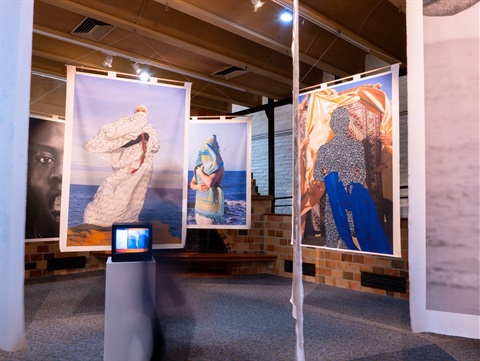 A photo of six photos printed on fabric hanging from the gallery ceiling, a plinth with a video work is in the centre of the room