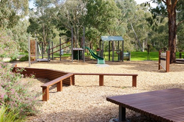 Wooden beams provide seating under a large gum tree, with a playground equipment surrounded by bright green grassy open space