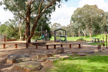 Wooden beams provide seating under a large gum tree, with a playground equipment surrounded by bright green grassy open space