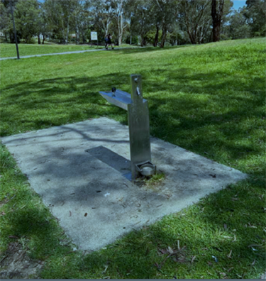 A rectangular concrete square sits in a grassed area, there is a drinking fountain and tap filler on the concrete