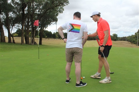 Photo of two people playing golf. One is wearing a tshirt with the rainbow pride flag on it.