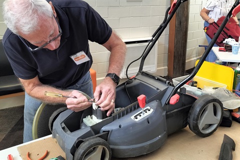 An older person is using a screwdriver on a lawnmower, there are tools on the bench next to the mower