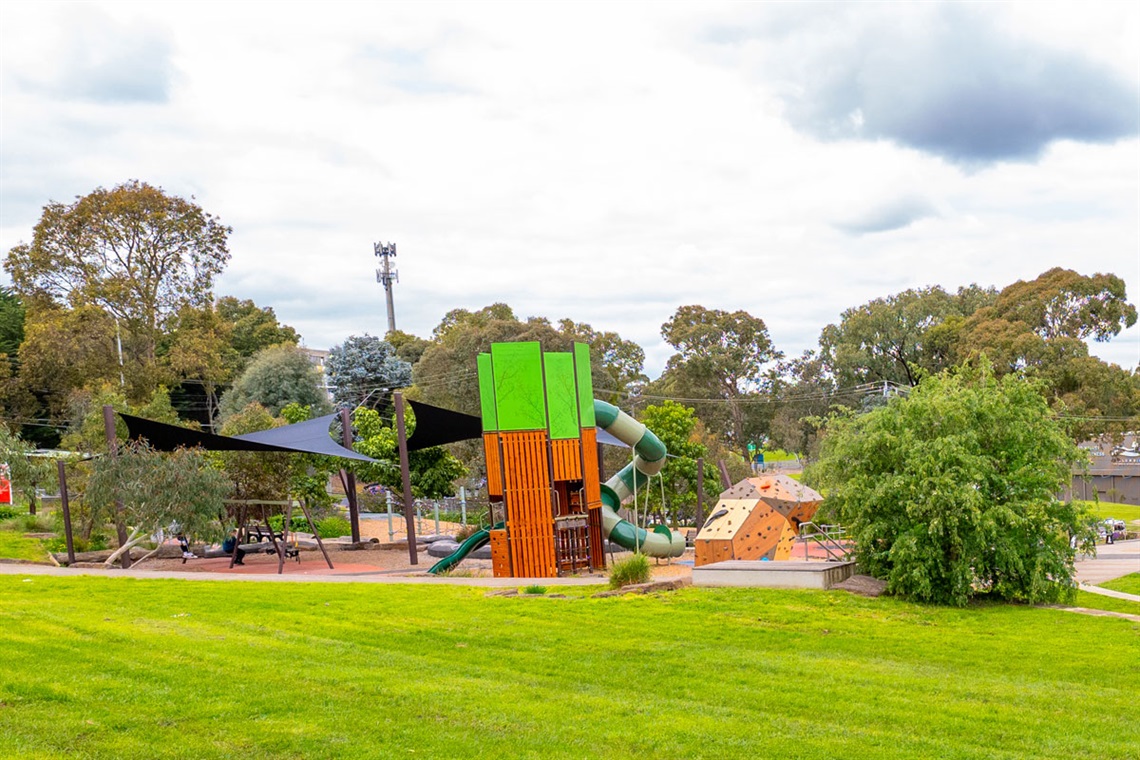 A brightly painted building sits behind a paved area surrounded by seating. There is a playground next to it with shade and large climbing structures.