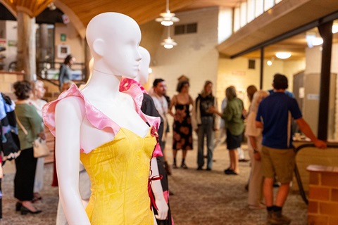 Photo of two mannequins in textile pieces (the front dress is yellow and pink) with audience members behind enjoying the opening.