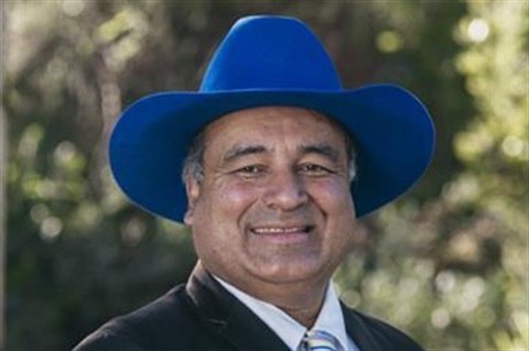 Photo of a man wearing a suit and tie and a large blue cowboy hat. He is smiling and there are trees behind him.